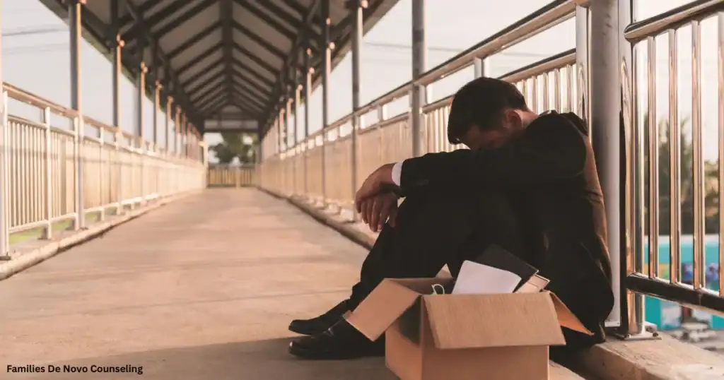 A businessman wearing a business suit sits alone next to a cardboard box along a covered walkway, demonstrating the emotional impact of losing structure during summer, emotional stress, change of life, and the need for mental health assistance.